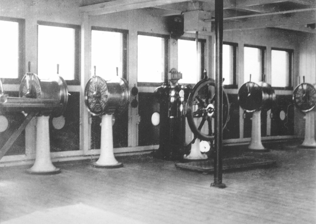 Olympic’s Navigating Bridge, showing the binnacle, wheel, and four of the five engine order telgraphs. The fold-down chart table can also be seen at the far left of the image. The dark objects below the windows are shutters that could be used if desire